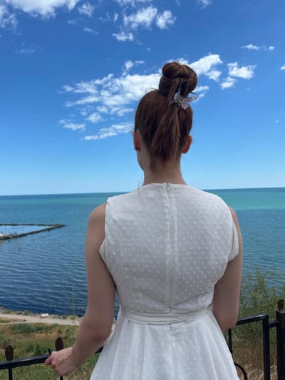The image depicts a young woman with reddish-brown hair pulled up in a bun, secured with a decorative butterfly clip. She is viewed from the back, wearing a white dress with a textured pattern. The woman is standing on a slight elevation, holding onto a black metal railing, and looking out at the sea. The sea is a deep blue-green, with a clear blue sky above dotted with white clouds. In the distance, a triangular-shaped structure extends into the water, with a few people visible on it. The coastline in the foreground is grassy and slightly uneven. The scene appears to be set in Kryzhanivka, Ukraine, a coastal village near Odesa.