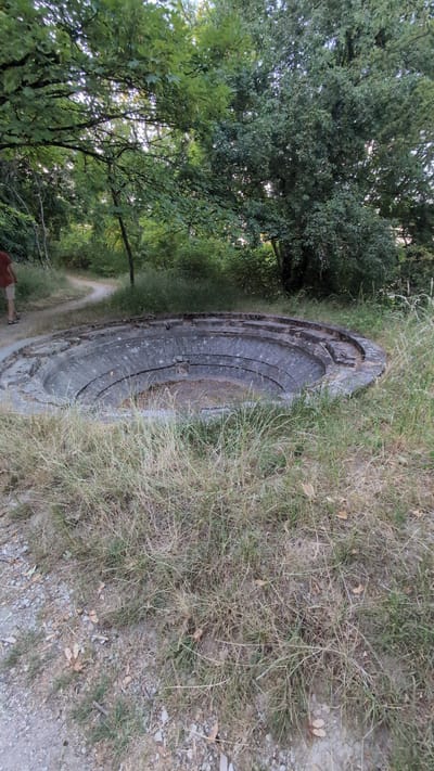 The image depicts a circular, tiered concrete structure, possibly a former well or cistern, situated in a grassy, wooded area. The structure is weathered and appears partially filled with debris. A dirt path winds behind it, and a person wearing a red shirt and khaki shorts is walking away from the camera on this path. The surrounding vegetation consists of tall, dry grass and lush green trees. The lighting suggests it is late afternoon or early evening. This structure is located in the city of Saint-Apollinaire, France, specifically in the vicinity of the former German artillery battery, which is now a protected historical site.