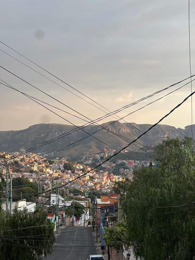 The image depicts an elevated panoramic view of Guanajuato City, Mexico, under a largely overcast sky with hints of pale light. The foreground features a downward-sloping, asphalt street flanked by buildings and verdant trees. Numerous black electrical and communication lines crisscross the upper two-thirds of the frame, converging and diverging. A white vehicle is partially visible at the bottom of the street. In the midground, the city unfolds with a dense arrangement of residential and commercial structures, notable for their varied and vibrant color palette, including shades of orange, pink, yellow, blue, and green, built into the natural hillside terrain. The background is dominated by large, rugged mountains covered in sparse vegetation, rising against the pale sky. No people, specific actions, or interactions are discernible within the frame.