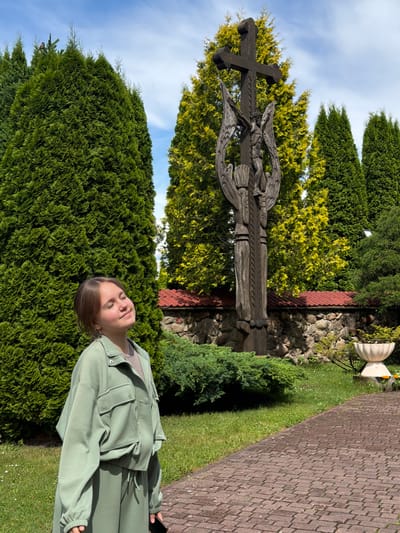 In a sunlit outdoor scene, a young girl with her eyes closed and a serene expression stands in front of a tall wooden cross featuring a carved crucifixion and angelic figures. She is wearing a light green tracksuit and jacket. Behind the cross, a stone wall with a red-tiled roof runs horizontally, and lush green and yellow coniferous trees form a backdrop under a bright blue sky with scattered clouds. The foreground features a paved brick pathway and a well-maintained lawn with some green shrubbery. The location is identified as Гервяцкі сельскі Савет, Belarus.