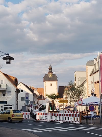 The image depicts a street scene in Obernburg am Main, Germany, under a partially cloudy sky with evident late-day sunlight illuminating buildings from the right. In the foreground, a multi-lane asphalt road features a white pedestrian crossing. A series of white and red striped construction barriers block the central section of the road.
On the left, a yellow Volkswagen Polo, bearing a German license plate (LJP-partially visible), is positioned on the road, partially obscuring additional barriers. Further left, multi-story buildings with white facades and reddish-brown tiled roofs line the street. A black, lantern-style streetlamp is visible in the upper-left. Traffic signs, including a circular "20 km/h" speed limit and blue directional signs, are affixed to posts and a building on the left.
The midground features the prominent Altes Rathaus (Old Town Hall) tower, a white structure with a dark, conical roof and a gold-faced clock, centrally positioned. A small palm tree stands near its base. A white van with a visible driver is positioned behind the construction barriers near the tower. A yellow "Umleitung" (Detour) sign points right.
To the right, a white tent branded "INTERSPORT" covers an outdoor dining or seating area where multiple individuals are seated at tables. Adjacent traffic signs include a red circular "no entry for all vehicles" sign and a blue circular "no stopping/parking" sign. Buildings with white facades and grey curved roofs are visible beyond the tent. The scene indicates temporary road modifications or an event.
