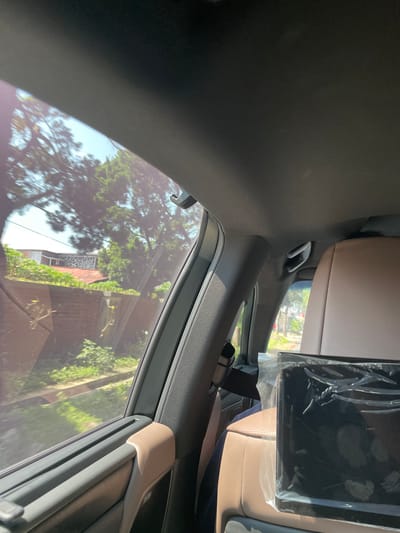 The image displays the interior of a moving vehicle and the view through its rear left passenger window, set in Kayuambon, Indonesia. From an interior perspective, likely from the front passenger area looking towards the rear left, the vehicle's dark gray headliner and a section of the B-pillar are visible. A beige grab handle is affixed to the B-pillar, and another is present further forward on the headliner. On the right, the back of a light brown, perforated leather front passenger seat is prominent, with a black, rectangular electronic device, encased in clear plastic wrapping, mounted on its surface.

Through the window, a sunlit exterior environment is observed, characterized by lush green foliage including dense trees and bushes, with slight motion blur indicating vehicle movement. In the background, residential structures are discernible; one building features a reddish-brown roof, and another exhibits a distinctive patterned upper section. A brick wall runs along the bottom of the visible structures, partially obscured by vegetation. Thin power lines are visible against patches of clear blue sky above the trees.