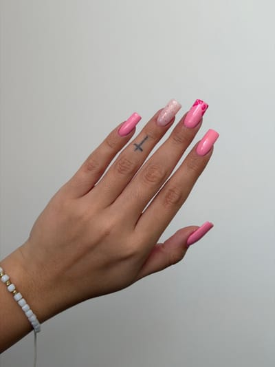 A human hand with pink acrylic nails is positioned against a plain white background. The nails are squared off with rounded corners and feature different designs. The index finger nail has a pink base with darker pink hearts. The middle finger nail is a speckled light pink and cream color. The ring finger and pinky finger nails are solid pink. A small tattoo of a cross is visible on the side of the ring finger. A bracelet of white and gold beads is around the wrist. No location or people besides the hand are apparent.