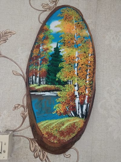 The image shows a piece of wall art, likely made of sand or small colorful pebbles, depicting an autumn landscape with birch trees and a body of water. The artwork is oval-shaped and mounted on a wall with a patterned wallpaper. To the left of the artwork, a light switch is visible. The location is identified as Kürşım, Kazakhstan.