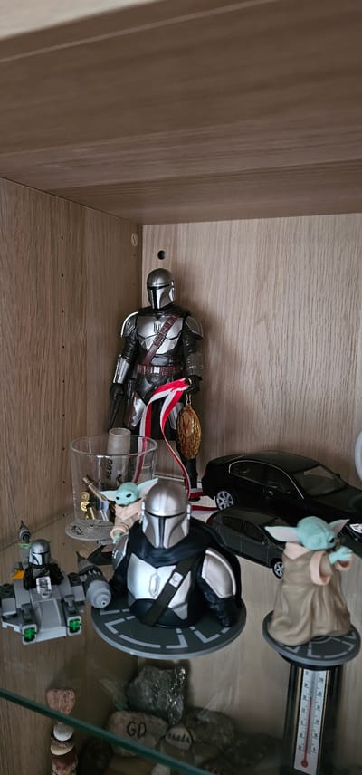 This image depicts a shelf within a wooden cabinet, likely in a home in Pabianice, Poland. The shelf is populated with various Star Wars-themed collectibles. Prominently featured is a Mandalorian action figure, standing tall and wearing its iconic silver armor. A medal on a red and white ribbon hangs from its hand. Below the Mandalorian, a small clear glass contains what appear to be bullet casings and a rolled-up paper tube. A Baby Yoda figure is also visible next to the glass. Further down the shelf, two more Baby Yoda figures are present, one near a small Lego-like vehicle and another on a stand next to a thermometer. Two miniature black cars are also on the shelf, one larger sedan and a smaller hatchback. The background of the shelf is the natural wood grain of the cabinet.