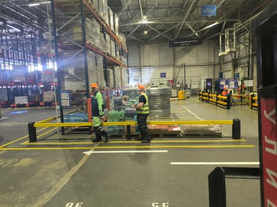 The image depicts a warehouse interior, specifically within the city of Perepechino, Russia, as indicated by signage. Two male workers, dressed in high-visibility vests and work pants, are the primary subjects. One worker, on the left, wears an orange hard hat and a green and blue vest, walking away from the camera. The second worker, on the right, also wears an orange hard hat and a yellow vest, and is seen carrying a case of bottled water. They are walking through a designated aisle marked by yellow lines on the floor, with a yellow safety barrier to their right.

The environment is a large industrial space with towering metal shelving units stacked high with pallets of goods, many of which are shrink-wrapped. The ceiling features exposed metal beams and lighting fixtures. Various pieces of industrial equipment, including what appears to be a pallet jack with a yellow gas cylinder, are visible in the background. Signage in Russian is present, with one prominent black sign reading "ЗОНА СОЗДАНИЯ ЦЕННОСТИ ПЕРЕУПАКОВКА" (Value Creation Zone Repackaging) and another blue sign reading "ЗОНА ПЕРЕУПАКОВКИ И ВОЗВРАТА" (Repackaging and Returns Zone). The floor is asphalt or concrete, with white directional arrows painted on it. The overall scene conveys a busy operational warehouse environment.