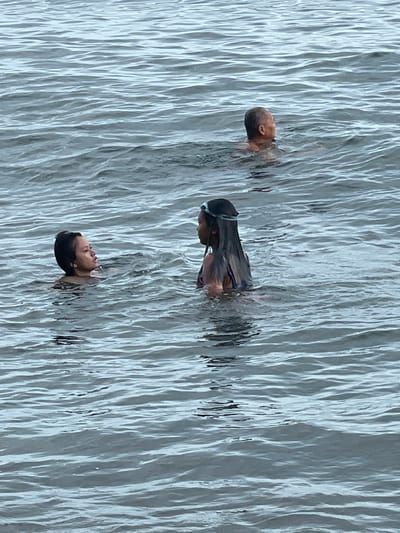 In the calm, rippling waters off Dauin, Philippines, three individuals are visible. In the background, a person with a shaved head is swimming, facing to the right. In the mid-ground, a person with dark hair and swimming goggles on their head is in the water, facing left and engaged in conversation with another person. This second person, in the mid-ground to the left, has dark hair and is also swimming, looking towards the person with goggles. The environment is an open body of water with moderate waves.
