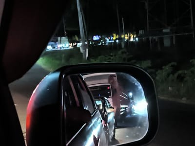 The image shows a view from inside a car, looking at the side mirror on a dark night. The mirror reflects a bus and a car following behind. The location appears to be Keudetanjong, Indonesia, based on other images taken around this location. In the background, a streetscape with various lights suggests a populated area. A transmission tower is faintly visible to the right. Greenery borders the road on the right side of the image.