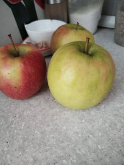 The image shows three apples on a speckled countertop. One apple in the foreground on the left is predominantly red with some yellow and green. Behind it and slightly to the right is another apple that is mostly green with some reddish blush. In the background, the third apple is also primarily green with red tones. To the left of the apples, a white bowl with some garlic cloves inside is visible. In the far background, blurred kitchen items are present, including what appears to be a canister of sugar and another container with a darker substance. The lighting suggests an indoor setting, likely a kitchen, during the daytime. There is no visible text in the image.