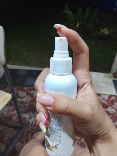 This media file features a close-up shot of a person's hand holding a white spray bottle. The hand, adorned with colorful French manicured nails featuring pink, yellow, and a touch of bright pink and green accents, is positioned to grasp the bottle's spray top. The bottle itself is white with a ribbed cap and a dispenser nozzle visible at the top. The label on the bottle contains text in what appears to be a non-Latin script, with some visible illustrations of snails.
The background is softly blurred, indicating an outdoor setting. Green foliage and plants are visible, suggesting a garden or natural environment. A hint of a patterned rug and a chair in the lower left corner further ground the scene in an outdoor living space. The lighting suggests it is either dusk or evening, with a dark sky and ambient light illuminating the hand and bottle. The overall mood is casual and domestic. The text on the bottle, partially visible, includes the phrase "СПИЙ МЛРАЛДАУШЫ" and mentions "мугополис" and "каразда," along with what appears to be a product code and a year "2014".