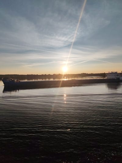 A wide shot captures a barge sailing on a calm river during sunset. The sun, low on the horizon, casts a golden glow across the sky, illuminating contrails from airplanes. The water's surface reflects the light and the sky, creating shimmering patterns. The barge, a long, flat-bottomed vessel, is dark against the bright water. The background features a silhouetted treeline and a hint of buildings on the distant shore, suggesting a location near a town or village. The scene is serene and peaceful, evoking a sense of tranquility. The location is identified as Bol'shoi Dvor, Russia.