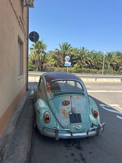 A rusty, light blue Volkswagen Beetle is parked on the side of a street in Muravera, Italy, beside a building with a pale yellow facade. The car, showing significant signs of wear with a heavily rusted roof and faded paint, faces away from the camera. Its rear bumper is chrome, and a license plate with the characters "GE58 5021" is visible. The street is paved, with white lane markings on the right. Behind the car, a line of palm trees stretches across the background, suggesting a Mediterranean climate. A guardrail separates the street from a road in the distance. There are traffic signs present, including a blue directional sign with white lettering indicating "Via Sarrabus." The sky is clear and blue.