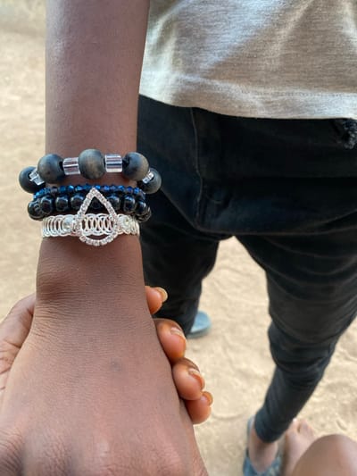 The scene is set outdoors on sandy, light-brown ground in Idiroko, Nigeria. The image focuses on two individuals, primarily showing their hands and lower bodies. In the lower left foreground, a dark-skinned hand, with fingernails exhibiting an orange-brown tint, firmly grasps the lower forearm of a second individual. This second individual, also dark-skinned, wears a stack of four bracelets on their forearm: the top bracelet features large dark beads separated by clear cylindrical beads; the second consists of small, faceted dark blue beads; the third is composed of small, dark, spherical beads; and the bottom bracelet is a wide, silver-toned cuff with a braided pattern, adorned with a teardrop-shaped element encrusted with clear stones. The second individual's attire consists of a light-colored (likely grey or off-white) t-shirt and dark-colored (likely black) pants. Their feet are visible on the ground; one foot wears a blue sandal, and the other appears bare or minimally covered.