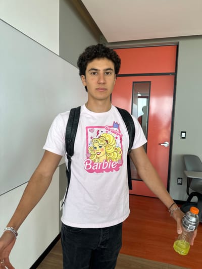 An eye-level photograph depicts a young adult male standing indoors against a two-toned wall and an orange door. The subject has dark, curly hair and is looking directly at the camera with a neutral expression. He is wearing a light pink short-sleeved t-shirt featuring a central graphic of the "Barbie" doll logo with blonde hair, accompanied by the text "Be unique." He wears dark-colored trousers and a black backpack over both shoulders. Multiple silver-colored bracelets and a white band are visible on his left wrist. His right wrist displays a dark-colored braided bracelet and a silver-colored chain bracelet. In his right hand, he holds a partially full, clear plastic bottle containing a yellow-green liquid, topped with a grey cap and an orange spout. The background includes a plain white wall on the left, transitioning to a grey section overhead. To the right, an orange door with a narrow vertical window is visible, reflecting a distorted image of a person. A grey wall panel is adjacent to the door. The floor is a solid orange color. A portion of a grey chair is visible in the lower right corner of the frame. This scene is located in Fraccionamiento Los Heroes Queretaro, Mexico.