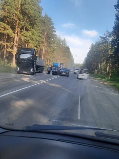 This media file captures a traffic jam on a road bordered by a dense forest on a sunny day, likely in the afternoon. The scene appears to be in Баюновские Ключи, Russia.
The primary subjects are several vehicles, including a black Volvo truck with "GLOBETROTTER" written on its side and license plate "E 890 TE 777". Other vehicles visible are cars and trucks, some appearing to be stopped or moving slowly. In the background, a white van and other vehicles are also part of the congestion. A few people are visible standing near the side of the road, potentially looking at the traffic or waiting.
The road itself is asphalt, with a white line marking a lane. There are signs of recent rain, with the road appearing wet and some mist or spray rising from the tires of the vehicles. The weather is sunny with blue skies and scattered clouds, suggesting a warm day. The dense pine forest on either side of the road indicates a rural or semi-rural setting.
The overall impression is one of a temporary disruption to travel, with the natural beauty of the forest contrasting with the man-made element of the traffic. There are no strong emotional cues conveyed, but the scene suggests a common experience of being stuck in traffic.
Visible text includes "GLOBETROTTER" on the truck, and license plates. A partial logo that may read "OZON" is visible on the side of a blue truck.