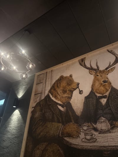 An upward-angled photograph depicts an interior scene in Le Grand-Saconnex, Switzerland, dominated by a large, framed artwork on the right side. The artwork features two anthropomorphic animals in a seated composition: a brown bear on the left, wearing a dark suit and waistcoat, with a pipe clenched in its mouth; and a male deer (stag) on the right, also attired in a dark suit, waistcoat, and bow tie. Between the two figures, a teapot and two stacked teacups with saucers are visible on a table.
To the left of the artwork, a textured dark grey wall is visible, with a black cylindrical wall sconce casting two distinct conical light patterns, one upward and one downward. Above the wall and artwork, a dark grey ceiling with diagonal ribbed panels extends across the upper half of the image. A modern chandelier hangs from the ceiling, composed of multiple clear glass spherical light fixtures arranged on a curved silver-toned frame. A circular dark-colored device is mounted on the ceiling.