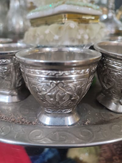 In the city of Blang Cut, Indonesia, a close-up shot focuses on a silver tray holding several intricately decorated silver cups. The cups are small and appear to be of traditional design, with detailed patterns etched into their surfaces. Some of the cups have a slightly raised rim and a pedestal base. The tray itself is also silver and exhibits decorative etchings along its edge, featuring swirling motifs. In the foreground, scattered on the tray around the cups, are small, granular particles, possibly spices or coffee grounds. The background is blurred, but suggests a decorative setting with hints of other metallic objects and possibly some floral elements, all rendered with soft lighting that creates a warm, inviting atmosphere. The overall impression is one of elegant presentation, likely for serving beverages or as decorative pieces.