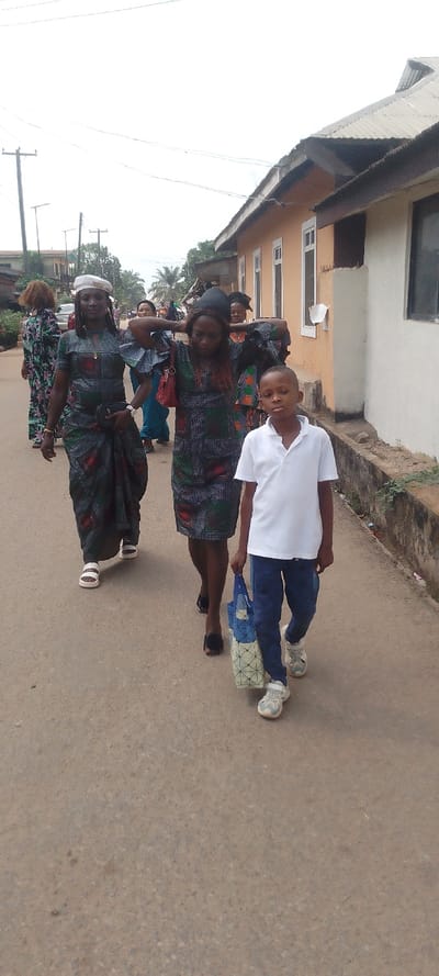 This daytime image captures a group of people, predominantly women, and a young boy, walking on a paved road in Ohafia, Nigeria, under an overcast sky.
In the foreground on the left, a woman is seen wearing a long, patterned traditional dress or wrap, a light-colored beret, and white sandals. She has a small black bag at her waist. Next to her, another woman wears a shorter, similarly patterned dress and dark open-toed shoes, with a red purse slung over her left shoulder. Both women look forward, with neutral to slightly smiling expressions.
To their right and slightly ahead, a young boy walks towards the viewer. He is dressed in a white polo shirt, dark blue trousers, and light-colored sneakers, holding a blue and white patterned reusable bag in his right hand. His gaze is directed towards the camera with a neutral expression.
Several other women, also adorned in patterned traditional attire, are visible in the background, suggesting they are part of the same procession or group.
The setting includes a single-story building with an orange/brown facade and a corrugated metal roof on the right side of the road. Utility poles and overhead wires are visible on the left, along with some distant buildings and vegetation, consistent with a residential or semi-urban area. The road surface is a light brown, possibly asphalt or compacted earth. The group appears to be walking together, likely to or from a communal gathering or event, given their matching traditional clothing. No discernible text is visible.
