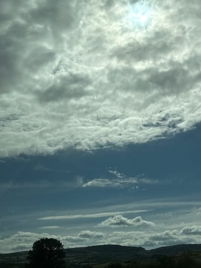 The image shows a landscape scene in Blajievo, Bulgaria, dominated by a dramatic sky. Large, thick clouds fill the upper portion of the frame, with the sun a bright, diffused light source peeking through them. Below the heavy cloud cover, patches of clear blue sky are visible. Further down, the sky transitions to a deeper blue as it meets a distant, undulating landscape of hills. A solitary, dark, bushy tree stands in silhouette against the lighter background in the lower left quadrant of the image. The hills are covered in vegetation, suggesting a rural or natural setting. The overall impression is one of a dynamic, possibly overcast sky over a serene, natural landscape.