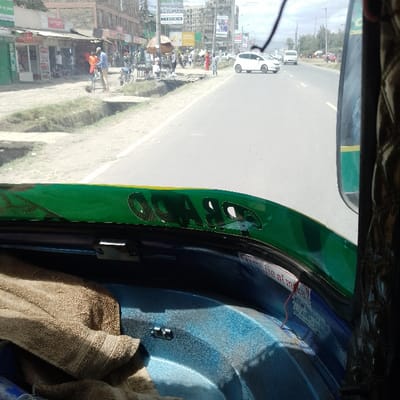 This image captures a daytime street scene in Ongata Rongai ward, Kenya, viewed from the interior of a vehicle. The immediate foreground displays the vehicle's dark blue interior trim, topped by a green and yellow band with partially visible black lettering, likely "RAO" or "RARO." A tan folded towel rests on the blue trim to the left, and a sticker with indecipherable text, from which a thin red string hangs, is affixed to the right side of the trim. A dark, possibly woven, material section and a black mirror mount are visible on the far right.

Beyond the vehicle's windshield, a two-lane asphalt road extends into the midground and background under a partly cloudy sky. A white hatchback or small SUV is positioned in the right lane, with a white van visible further down the road. The left side of the road features a dirt shoulder with a distinct drainage ditch. Beyond the ditch, a commercial street is active with multi-story buildings housing ground-floor businesses. Multiple individuals are visible: one man in an orange shirt and blue pants stands next to a bicycle on the dirt shoulder, while other pedestrians are dispersed on the sidewalk and near the buildings. An open brown umbrella or parasol stands on the left, near a small gathering of people, possibly indicating a vendor stall. Overhead power lines, signs, and advertisements are present along the street and buildings. Sparse vegetation grows in the ditch and along the roadside.