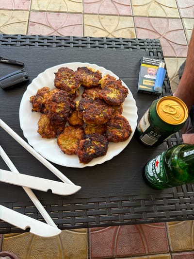 The scene is an outdoor setting, specifically a patio, located in the vicinity of Dragor, Bulgaria. A dark, woven-textured outdoor table is centrally featured. On the table, a white, scallop-edged plate is laden with approximately two dozen golden-brown, pan-fried patties or fritters, exhibiting visible green specks consistent with herbs or vegetables. To the right of the plate, a green aluminum can with a gold top and a green glass bottle of Heineken beer are positioned. Adjacent to these beverages, a pack of Dunhill Select cigarettes, displaying a health warning in Cyrillic ("Децата на пушачите е по-вероятно също да станат пушачи" - "Children of smokers are also more likely to become smokers") with an image of a baby holding a pacifier, stands next to a light blue disposable lighter. To the left of the plate, a black electronic device, possibly a small camera or key fob, with a strap bearing the text "maxwell" or "maxuell," is visible. Two white, flat, notched objects, possibly components of hangers or specialized utensils, are crossed on the table in the foreground. A partial view of a tanned, hairy right leg, wearing dark shorts, is visible on the far right side of the frame, resting near the table. The ground beneath the table is paved with multi-colored (pink, yellow, beige, terracotta) patterned patio tiles.