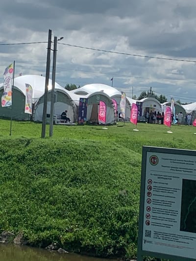 In the city of Arbuzov-Baran, Russia, a large, outdoor event is underway. Multiple white, geodesic-shaped tents are set up on a grassy field, suggesting a festival or exhibition. Colorful flags and banners are displayed prominently, some with logos and what appear to be numbering. In the foreground, a wooden sign with text and icons is partially visible, offering information or regulations about the area. The sky is overcast with dramatic clouds. Several people are visible in the background, some milling about near the tents, while one individual sits on a bench inside one of the tents, appearing to be reading. The overall scene depicts a community gathering in a semi-rural or park-like setting.