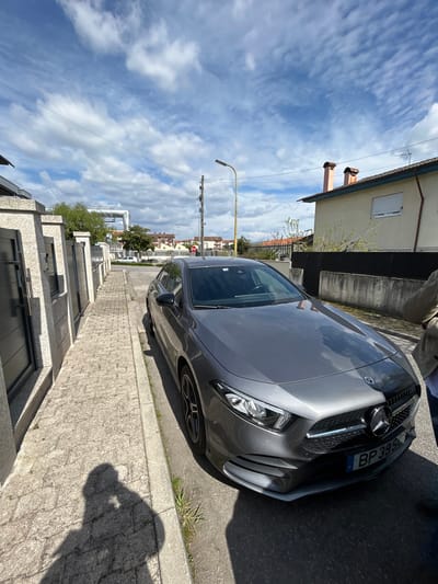The image displays a dark grey Mercedes-Benz sedan, license plate BP 39 SL, parked curbside on an asphalt street in Chaves, Portugal. The vehicle is positioned parallel to a raised cobblestone sidewalk on the left, which is bordered by a series of grey concrete pillars and dark metal gates leading to a residential property. Two distinct human shadows, likely the photographer and an accompanying individual, are cast onto the sidewalk and the street in the immediate foreground, with the lower torso and right leg of a person in light-colored attire visible at the far right edge next to the car.
On the opposite side of the street, to the right of the parked vehicle, a light-colored house with a red-tiled roof and two terracotta chimneys is visible, featuring a white roller shutter on a window. Further into the background, a yellow street light pole, a utility pole, and a partially obscured red octagonal stop sign mark a distant intersection. In the far distance, large, elevated grey pipes or industrial structures are discernible against a bright blue sky with scattered cumulus clouds. The scene is under direct sunlight, indicated by the sharp shadows cast, suggesting mid-morning to early afternoon conditions. Small patches of green vegetation are present along the street curb.