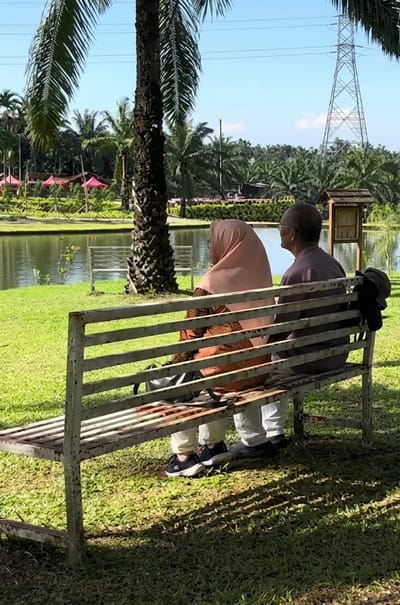 Two individuals are seated on a weathered, white metal bench in a grassy park area. The person on the left, viewed from behind, wears a light pink hijab and a patterned top. To their right, a person with a shaved head and glasses is wearing a plaid shirt and light-colored pants. They are both facing away from the camera, looking towards a body of water. Behind the bench, a large palm tree is prominent, and further in the background, a lake, more palm trees, and a high-voltage electricity pylon are visible under a clear blue sky. The area appears to be a recreational park in or around Bintang Meriah, Indonesia, suggested by the surrounding tropical vegetation.