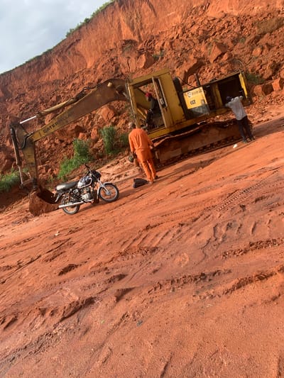 A construction site in Ijesha-Ijebu, Nigeria, features a large yellow excavator positioned on a dirt path. The ground is a reddish-brown dirt, with visible tire tracks and divots. Behind the excavator, a steep embankment of red earth and rocks rises sharply, topped with sparse green vegetation. In the foreground, a motorcycle is parked on the dirt path. Three individuals are present: one wearing orange clothing stands near the motorcycle, another person in a light-colored shirt is working on the rear of the excavator, and a third individual, wearing a red head covering, is seated in the excavator's cab. The sky above is overcast.