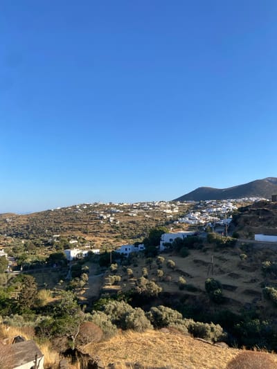 A daytime, elevated, wide-angle view depicts the village of Απολλωνία, Greece, situated across arid, terraced hillsides under a clear, unclouded blue sky. The landscape is characterized by extensive horizontal terracing, covered with dry, light-brown vegetation and scattered olive trees. Numerous white-washed, cubic buildings with flat roofs are distributed throughout the scene, forming clusters and individual structures on the slopes. A large, darker, potentially un-whitewashed structure occupies a ridge on the right horizon. No human subjects, animals, or active events are discernible.