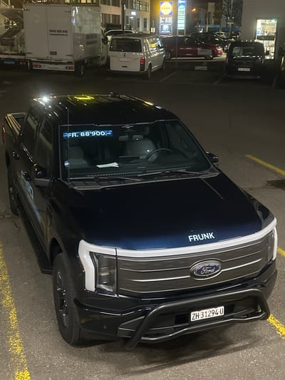 The image displays a dark blue Ford F-150 Lightning electric pickup truck parked in an illuminated commercial parking lot at night in Volketswil, Switzerland. The truck is angled, partially occupying a parking space delineated by faded yellow lines. The front of the vehicle features a black bull bar, a Ford emblem on the grille, and a Swiss license plate reading "ZH 31294 U". The word "FRUNK" is visible in white capital letters on the hood. On the driver's side, a "F-150 LIGHTNING" decal is present. An illuminated digital display within the windshield shows "FR. 88'900.-". The vehicle's interior appears unoccupied.
In the background, various other vehicles are parked. These include a white box truck with "0848 000 048 umzugszentrale.ch" text on its rear, a white passenger van, a red sedan, and a black passenger van. Building facades are visible behind the vehicles, featuring illuminated signs for "LIDL" and "UMB", confirming a commercial establishment setting consistent with areas in Volketswil. The parking lot surface is asphalt, reflecting ambient light from overhead fixtures. There are no individuals visible in the scene, and no actions or interactions are depicted.