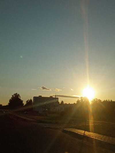 The image depicts a bright sunset casting a warm glow over a rural landscape. The sun, a brilliant orb, is positioned in the upper right quadrant, sending out lens flares and streaks of light across the sky. The sky itself transitions from a clear, pale blue on the left to a hazy yellow-orange near the sun. A few wispy clouds are visible, catching the light.
In the middle ground, silhouetted against the sunset, are trees and a building. The building appears to be a multi-story structure, possibly an apartment block or administrative building, with dark windows. The trees are dense and dark, their foliage obscuring details. A road or path runs alongside the building, and further to the right, a concrete structure, possibly a curb or wall, is visible. The ground area appears to be an open field or park.
The overall impression is one of a peaceful evening in a rural setting. The time of day is clearly sunset, indicated by the low angle of the sun and the warm light. The weather appears clear with a slight haze. The location cues, such as the building style and vegetation, suggest a European context. The image does not contain any visible text.