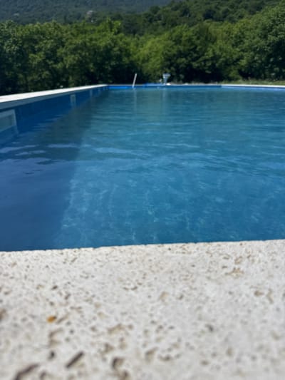 An outdoor swimming pool is shown from a low angle, with the textured edge of the pool in the foreground. The water is a vibrant blue, and the sun reflects off its surface, creating shimmering patterns. In the background, lush green trees line a hillside, with a hazy mountain range beyond. The scene is serene and suggests a peaceful, natural setting. The location is identified as Yenokavan, Armenia.