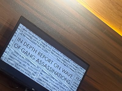 The image shows a television displaying the headline "In Depth Report On Wave of Gang Assassinations." The television is an LG model. The on-screen text suggests the article discusses gang-related killings that began two weeks prior, examined within the Metro section of a newspaper. The article appears to discuss the victims of the assassinations being associates of former Los Angeles crime lord Mickey Cohen. The television is sitting on a wooden surface, and the background is out of focus. The location of Eneka, Nigeria, is not evident in the image.