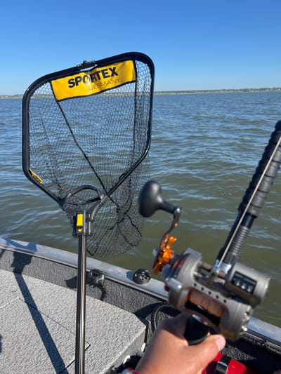 A person's hand holds a fishing reel on a boat. The reel is gray and silver with orange accents and a black handle. A fishing rod extends from the reel. In the background, a fishing net with a yellow banner that reads "SPORTEX GERMANY" is attached to a pole. The boat is on a body of water, likely a lake or river, under a clear blue sky. The far shore is visible in the distance, lined with trees and some buildings. This scene is not located in Novopichugovo, Russia, as there is no information in the image to suggest this location.