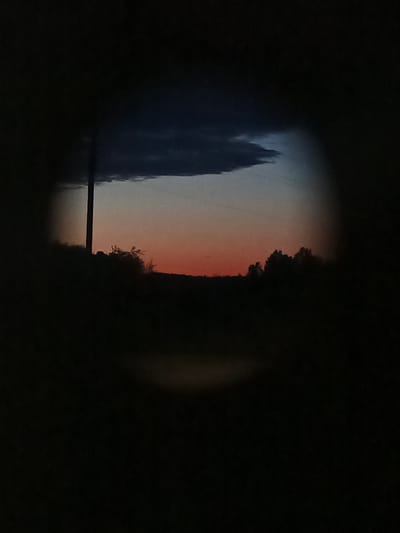 The image captures a twilight scene through a dark, vignetted frame, resembling a view through a telescope or binoculars. The sky displays a gradient of colors, transitioning from a deep, stormy gray at the top to a soft orange and pink hue near the horizon, indicative of sunset or sunrise. A dark, elongated cloud formation is prominent in the upper portion of the sky. In the mid-ground, silhouetted against the colorful sky, is a dark, undulating landscape, possibly hills or a treeline. A single, thin, vertical pole, likely a utility pole, stands prominently on the left side of the frame. The lower portion of the image is predominantly dark, with faint, shadowy outlines suggesting the presence of foliage or other terrestrial features. The overall impression is one of a rural or semi-rural setting at dusk, devoid of any human figures or explicit signs of urban activity. The location is identified as Воскресенское, Russia.