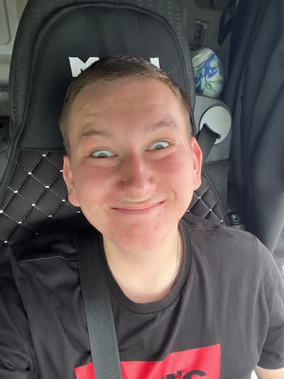 A close-up, high-angle selfie shows a young person with short brown hair and light blue eyes, looking directly at the camera with a wide, excited expression. Their face is flushed, and there are some red blemishes on their cheeks and chin. They are wearing a black t-shirt with a red graphic on it, and a silver chain necklace. The person is seated in a vehicle, as indicated by a black seat with white stitching and a seatbelt visible in the foreground. A dark curtain is in the background, and a small, decorative item with a floral pattern is visible to the right of the person's head. The overall scene is indoors and appears to be a personal, candid moment. The location of Старое Бобренево, Russia, is not evident from the visual information in the image.