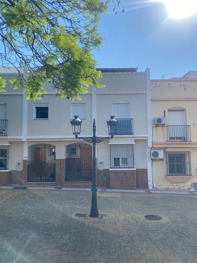 This is a photograph of a street in Cancelada, Spain, on a sunny day. A leafy tree branch with green foliage hangs into the frame from the upper left, partially obscuring the sky and the tops of buildings. The buildings are terraced houses with light-colored facades and cream-colored trim. Some of the buildings have balconies with black metal railings and barred windows. There are two air conditioning units visible on the facade of one of the buildings. In the foreground, a cobblestone street is visible, with chalk drawings on its surface. A black lamppost with two ornate lamps stands in the center of the street. The sky is clear and blue, with the sun glinting brightly in the upper right corner.