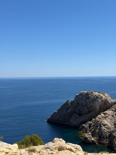 The image depicts a coastal scene at Cala Rajada, Spain. The right side of the frame features a rugged cliff face composed of gray rock and tan rock formations, with some greenery near the bottom edge. The cliff drops down to the blue water of the sea. In the distance, there is a white sail on the water. The sky occupies the left half of the frame and is a clear, gradient blue.