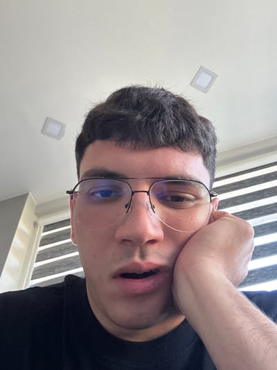 The image shows a close-up, low-angle selfie of a young man with dark hair and glasses. He is resting his right cheek on his hand, with his mouth slightly open and his eyes looking towards the camera. He is wearing a black t-shirt. The background consists of a white ceiling with two square lights, and a window with horizontal blinds. The setting appears to be indoors, possibly a home or office. There is no indication of the location being in the city of Dibich, Bulgaria.