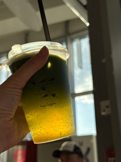 A hand holds a transparent plastic cup filled with a two-toned liquid, dark green at the bottom and yellow with bubbles at the top. The cup has a clear plastic lid and a black straw is partially visible. The label on the cup reads "Surf Coffee" next to a tic-tac-toe grid with coffee cup icons. The background shows a blurry exterior view through a window, with a grey structure and an indistinct building in the distance. The location is Κοщаково, Russia.