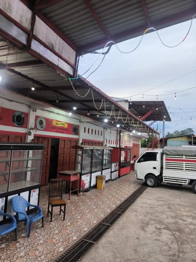 The image depicts an outdoor commercial area, likely a cafe or a row of small shops, in Sukamulia, Indonesia. A long, single-story building with white walls and distinctive red horizontal trim and borders forms the primary subject. Several dark wooden double doors are visible, some closed and one slightly ajar. Above one section, a yellow sign reads "BEK TEUWO CAFE ESPRESSO CEK SAM," with painted images of coffee cups adorning the red wall space.

The building is covered by a corrugated metal roof that extends forward, creating a wide awning over an outdoor patio area. Underneath this awning, numerous bare-bulb string lights and a wavy LED strip light are suspended, with some lights illuminated. The ground is paved with patterned tiles featuring circular designs in various earthy tones.

In the foreground and mid-ground, several empty glass-fronted display cases, likely for food or beverages, are arranged. A small wooden table, two blue plastic chairs, and one dark wooden chair are also present. A yellow bin is visible near the display cases. A metal drainage grate runs across the ground, separating the tiled patio from a concrete section. A white light commercial truck, possibly a pickup with a covered flatbed, is parked on the concrete pavement.

The sky appears overcast, suggesting either an early morning or late afternoon/evening setting, complemented by the illuminated lights. No people are visible, contributing to a quiet and still atmosphere.