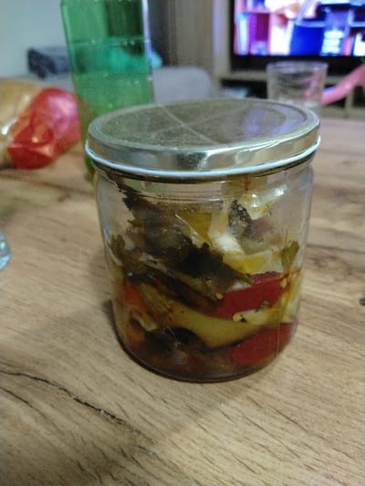 This image depicts a clear glass jar with a metallic, two-piece lid, containing various pickled vegetables including red, green, and yellow pieces, submerged in a liquid. The jar sits on a light brown wooden surface with visible grain and minor surface cracks. In the blurred midground, a tall, translucent green bottle or cup and a red, possibly plastic-wrapped object are visible. The heavily blurred background shows a television screen displaying colorful patterns, a clear drinking glass, and a pink object. The scene takes place indoors, in what appears to be a domestic setting within the city of Oreshene, Bulgaria. No individuals or specific actions are observed.
