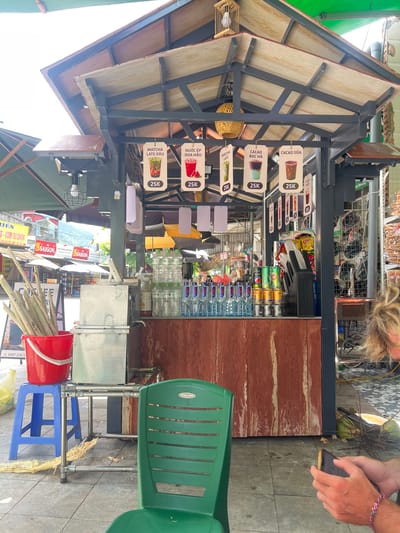 The image depicts a street-side beverage stall in Tân Quang Commune, Vietnam. The stall features a covered roof supported by metal beams. Hanging from the roof are several signs displaying drinks and their prices, with "25K" appearing on multiple signs. A green plastic chair is placed in front of the stall. To the left, a red bucket filled with sugarcane stalks sits on a blue plastic stool. In the background, shelves are stocked with various goods, including snacks and bottled drinks. The ground is paved with tiles. A person's hands, holding a mobile phone, are visible in the lower right corner of the frame. Coconuts are scattered near the base of the stall.