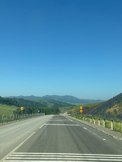 The image shows a view from a moving vehicle on a paved road. The road is divided into two lanes, with white lines marking the boundaries. There are speed limit signs visible on the side of the road, one indicating 30 km/h and another 50 km/h. The landscape is characterized by rolling green hills covered in trees and vegetation. In the distance, mountains are visible under a clear blue sky. There are no people or animals clearly visible in the frame. The scene appears to be in a rural or mountainous area, possibly along a highway.