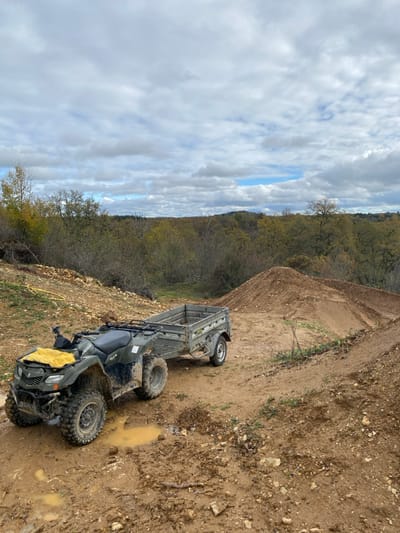 The scene, located in Saint-Denis-Catus, France, depicts an unpaved, rural landscape under an overcast sky with visible patches of blue. A mud-covered dark green Suzuki KingQuad all-terrain vehicle (ATV) is stationary on wet, rutted, reddish-brown soil. A yellow cloth is draped over the ATV's front utility rack. An empty, grey, single-axle utility trailer is hitched to the rear of the ATV. To the right, a large, freshly formed mound of reddish-brown soil is prominent, with another smaller earth mound visible further right. To the left, a slope of exposed, lighter-colored soil mixed with gravel features a coiled yellow garden hose. The immediate foreground ground is characterized by muddy puddles. The background consists of rolling hills covered by a dense forest displaying autumnal foliage. No individuals are present.