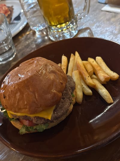 A burger and fries meal is presented on a dark brown plate, likely in a restaurant or dining establishment. Beside the plate, a glass mug filled with amber liquid, possibly beer, sits on the table. The burger features a glazed bun, a visible slice of cheese, a patty, and toppings such as lettuce and tomato. The fries are golden brown and appear to be seasoned. The environment seems to be a restaurant or bar, given the table setting and the drink. While I cannot definitively confirm the location as Tlaxcala City, Mexico, based solely on this image, the food presentation does not contradict such a setting.