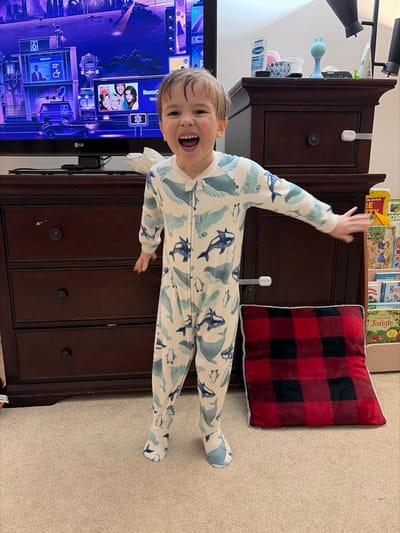 An indoor scene within Woodstock, Canada, depicts a young child standing centrally on a light brown carpeted floor. The child, appearing male, has light skin, short, damp-looking dark hair, and is wearing a full-body, footed pajama suit with a white background and repeating patterns of light blue and dark blue whales and penguins. He faces forward, smiling widely with an open mouth, revealing teeth, and his left arm is extended horizontally to the side with an open palm.

Behind the child, a dark brown wooden dresser or console table with three visible drawers and dark circular knobs spans the width of the frame. A white child safety latch is affixed to the upper right drawer. On the dresser's surface, a crumpled white tissue, a blue Cetaphil bottle, a pink-lidded container, and a blue, rabbit-eared toy are visible. Above the dresser, a large, black-framed flat-screen LG television displays a colorful animated cityscape with icons and text such as "Roku City," "6 News," and a promotional image for "Who's the Boss."

To the child's right, a red and black buffalo plaid cushion with a white furry edge rests on the carpet. Adjacent to the cushion, a small, light-colored wooden bookshelf filled with children's books is partially visible. The background consists of a plain white wall, with two black overhead fixtures or supports visible in the upper right corner.