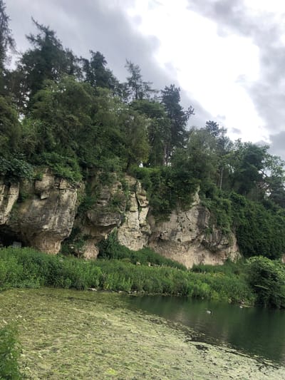 The image shows a rocky outcrop covered in greenery beside a body of water in Bolsover, United Kingdom. The sky is overcast. Trees grow atop the outcrop, and the water is algae-covered.