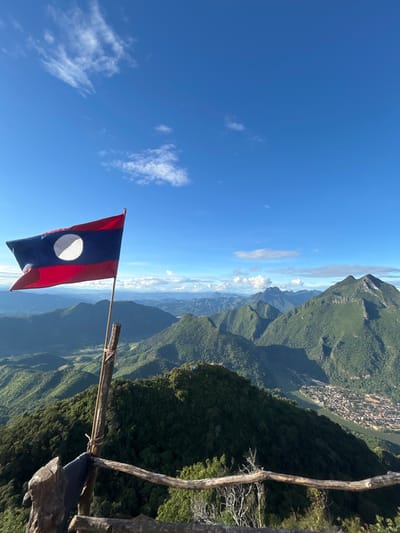 A forensic examination of the image reveals a high-angle panoramic view from a mountainous vantage point in Nongkhiao-Nua, Laos. In the mid-left foreground, the national flag of Laos, displaying red, blue, and red horizontal stripes with a central white disc, is attached to a bamboo pole. The pole is secured to a rough-hewn wooden post, which forms part of a rudimentary wooden railing or fence extending across the lower portion of the frame.
The background presents a vast, undulating landscape dominated by densely forested green mountains and peaks, receding into the distance under a clear, bright blue sky adorned with sparse white cumulus clouds. To the far right, nestled in a valley among the mountains, a town or village settlement, consistent with Nongkhiao-Nua, is visible alongside a winding body of water, likely the Nam Ou River. The structures within the settlement appear as concentrated clusters of buildings. No individuals are discernible within the scene. The lighting indicates natural daylight, casting distinct shadows on the mountainous terrain.